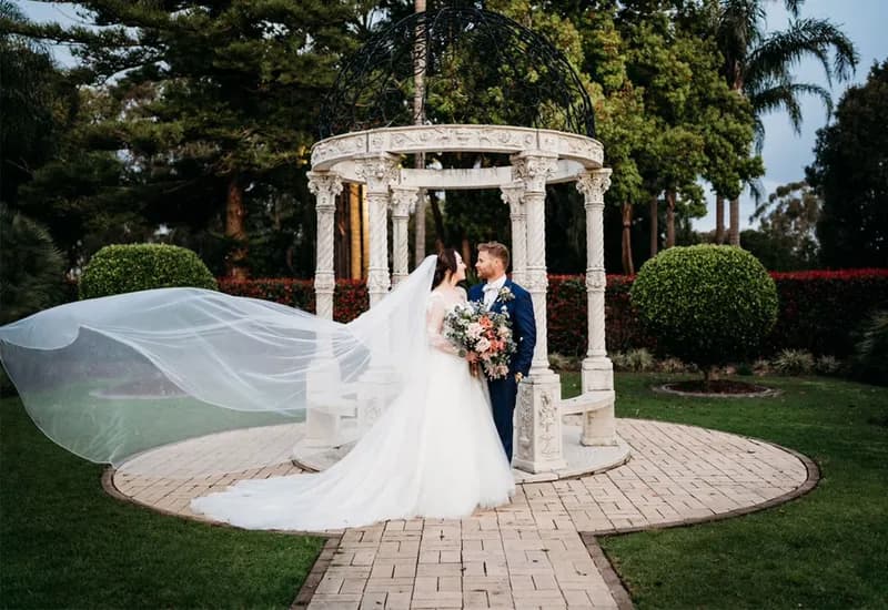 A bride and groom by a garden rotunda, her veil lifting in the breeze