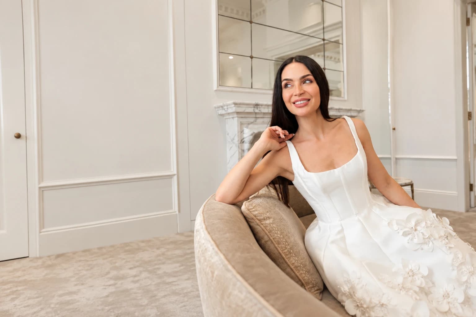 A bride in the Gia Flora mini dress by Steven Khalil, seated on a velvet sofa in a bridal salon: an ivory corset-seamed bodice on wide straps and a skirt covered in three-dimensional pearl-centred flowers