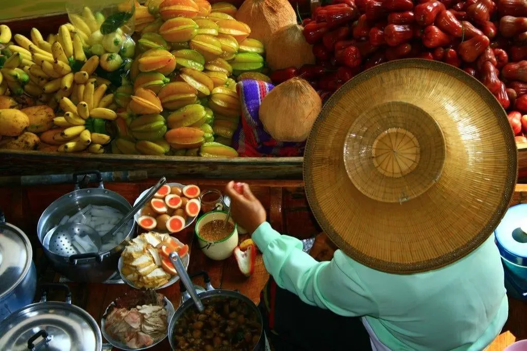 A vendor in a straw hat with fruit and bowls of food on a market boat