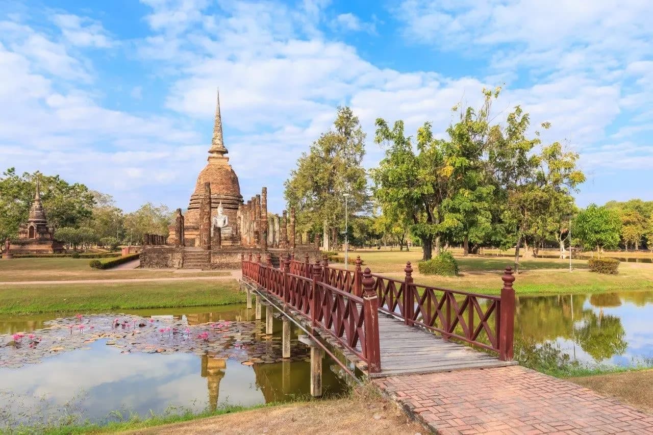 A red wooden footbridge crossing a pond towards a ruined chedi at Sukhothai