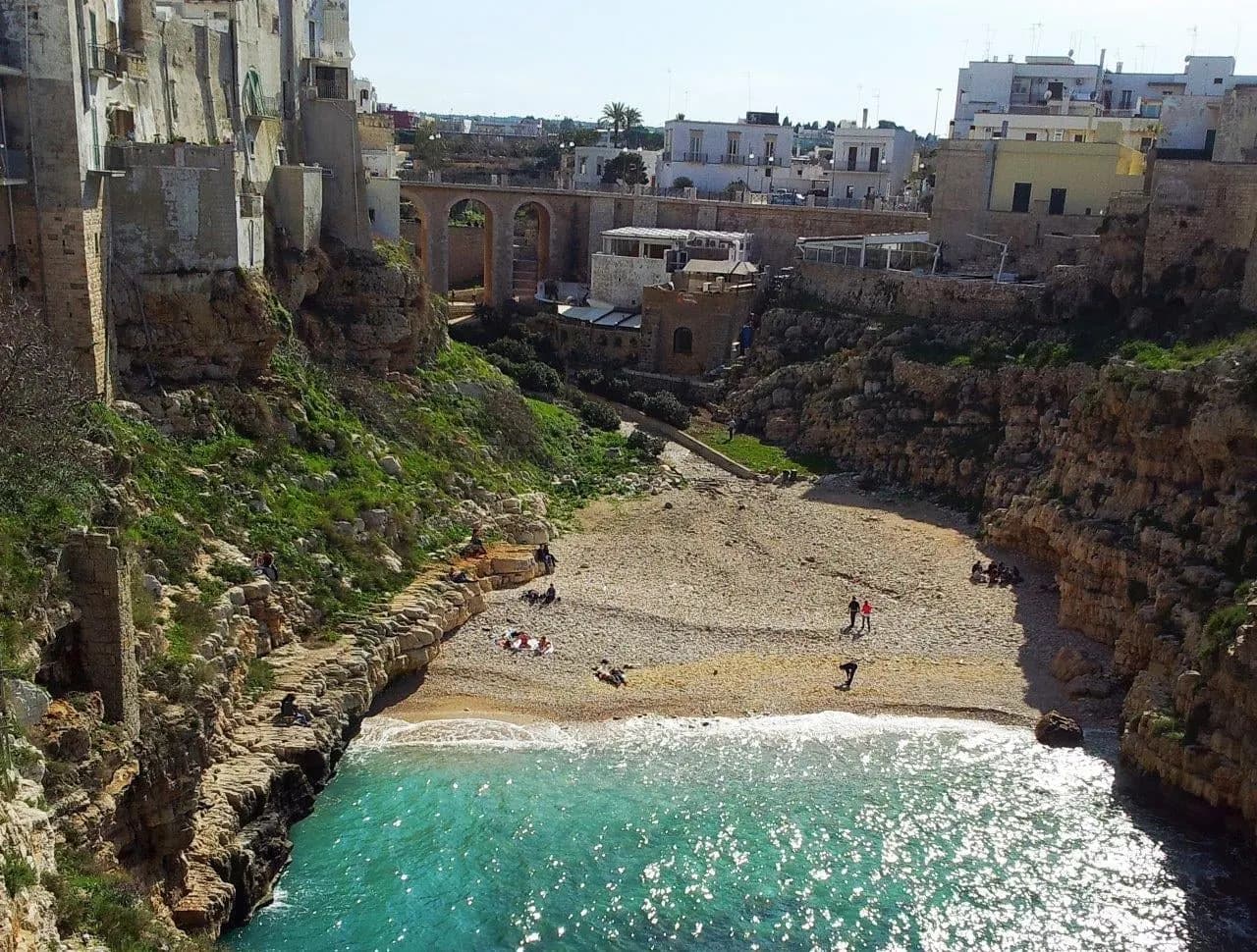 A small pebble cove with turquoise water below an old stone bridge and town
