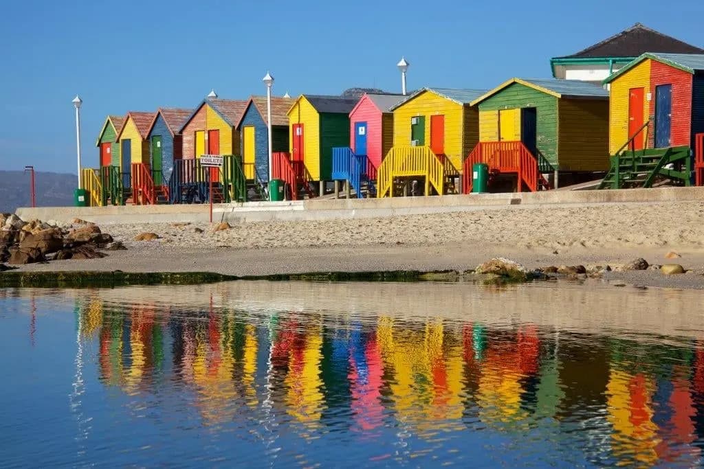 A row of brightly painted bathing huts reflected in still water on a Cape beach