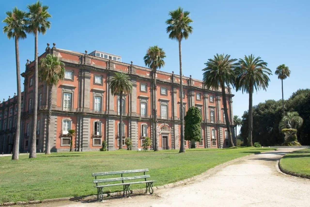 A red and grey palace behind tall palm trees and lawns in Naples