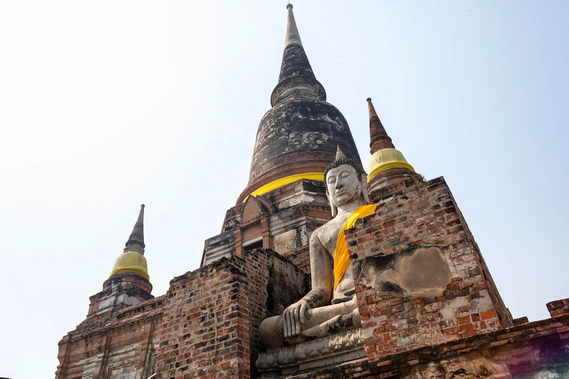 A seated Buddha in a saffron sash in front of a brick chedi in Ayutthaya