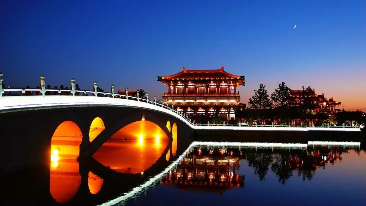 An illuminated arched bridge and Tang-style hall reflected in still water at dusk