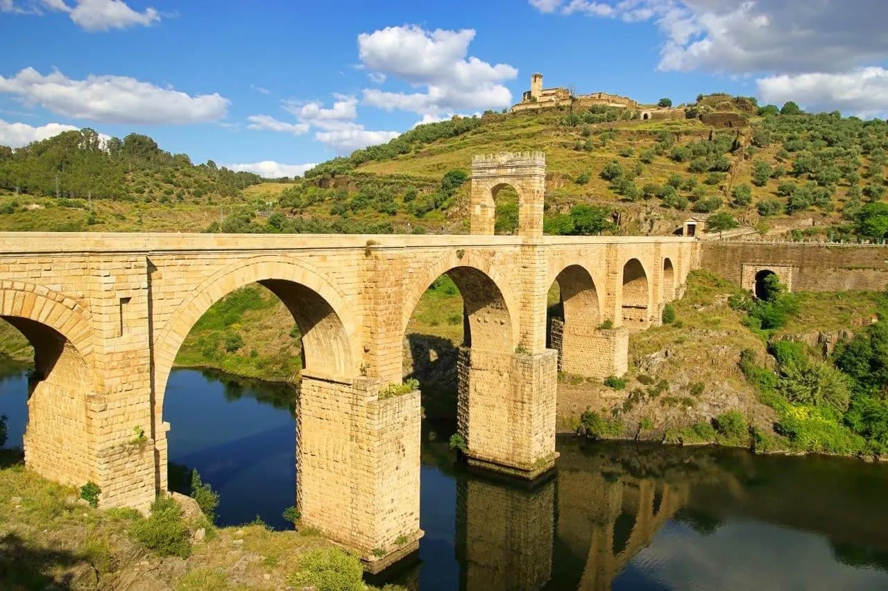 A tall Roman stone bridge reflected in the river below green hills