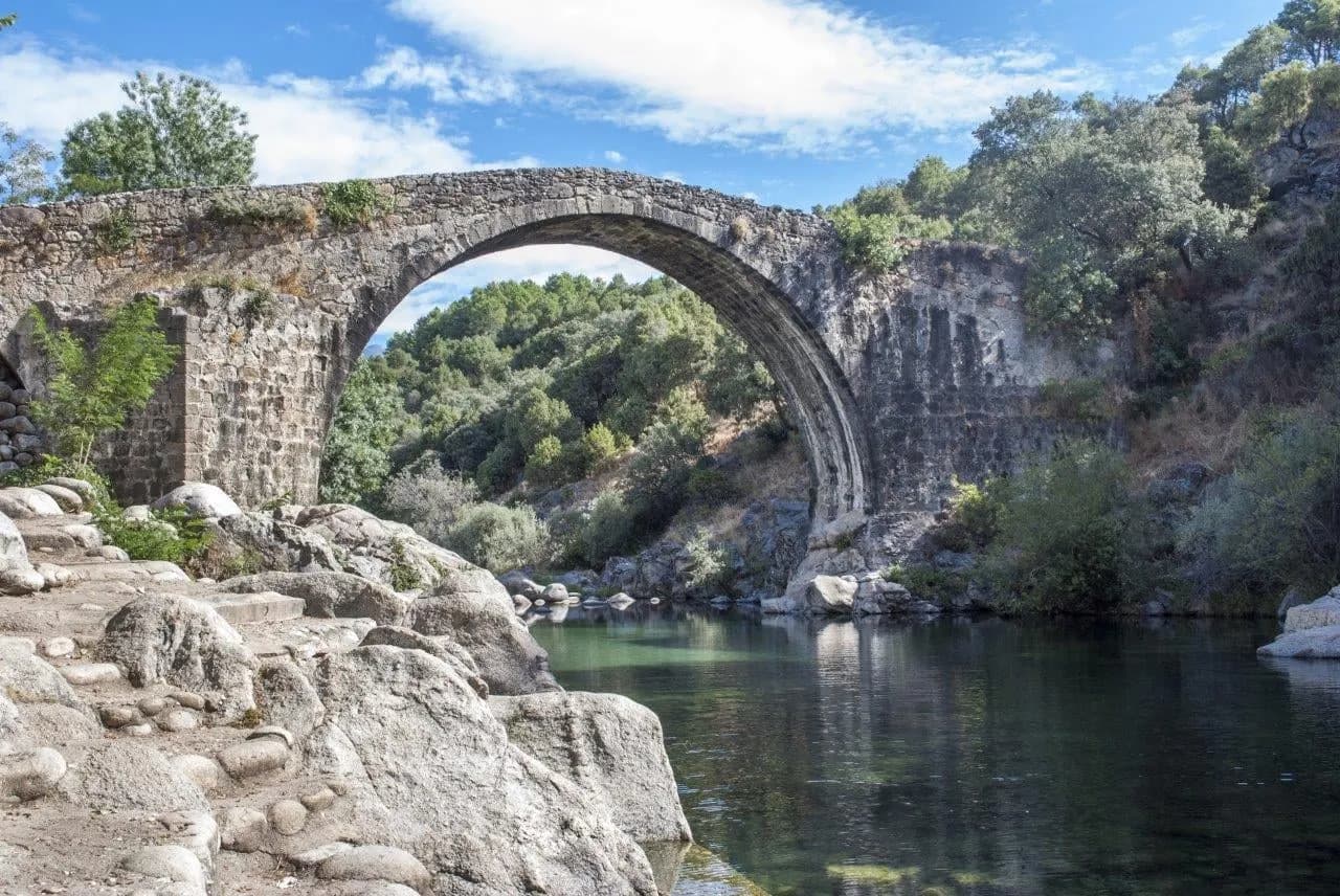 An old single-arch stone bridge over a clear river among rocks and trees