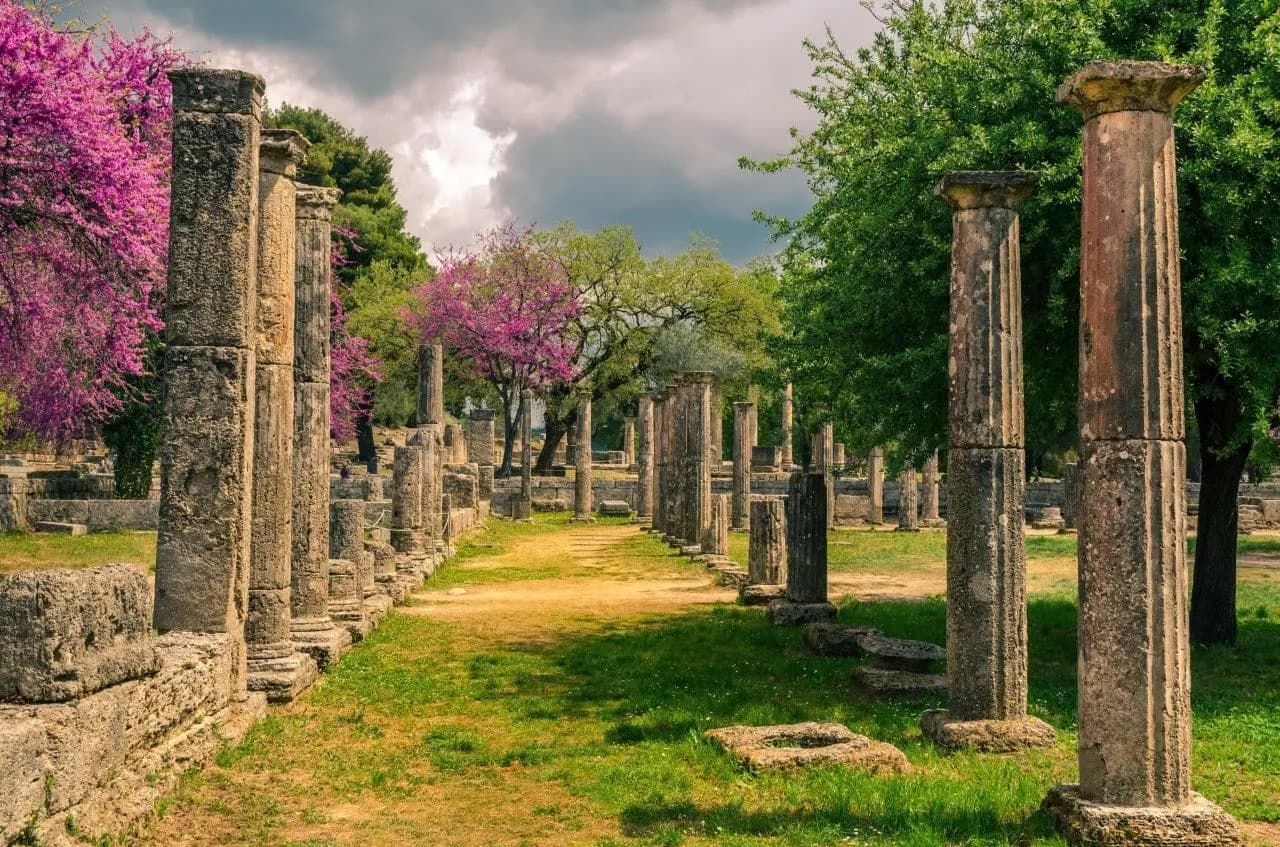 Columns lining a grassy path at ancient Olympia, with trees in pink blossom