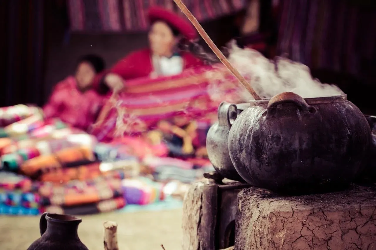 A clay pot steaming beside woven textiles, with a weaver at work behind