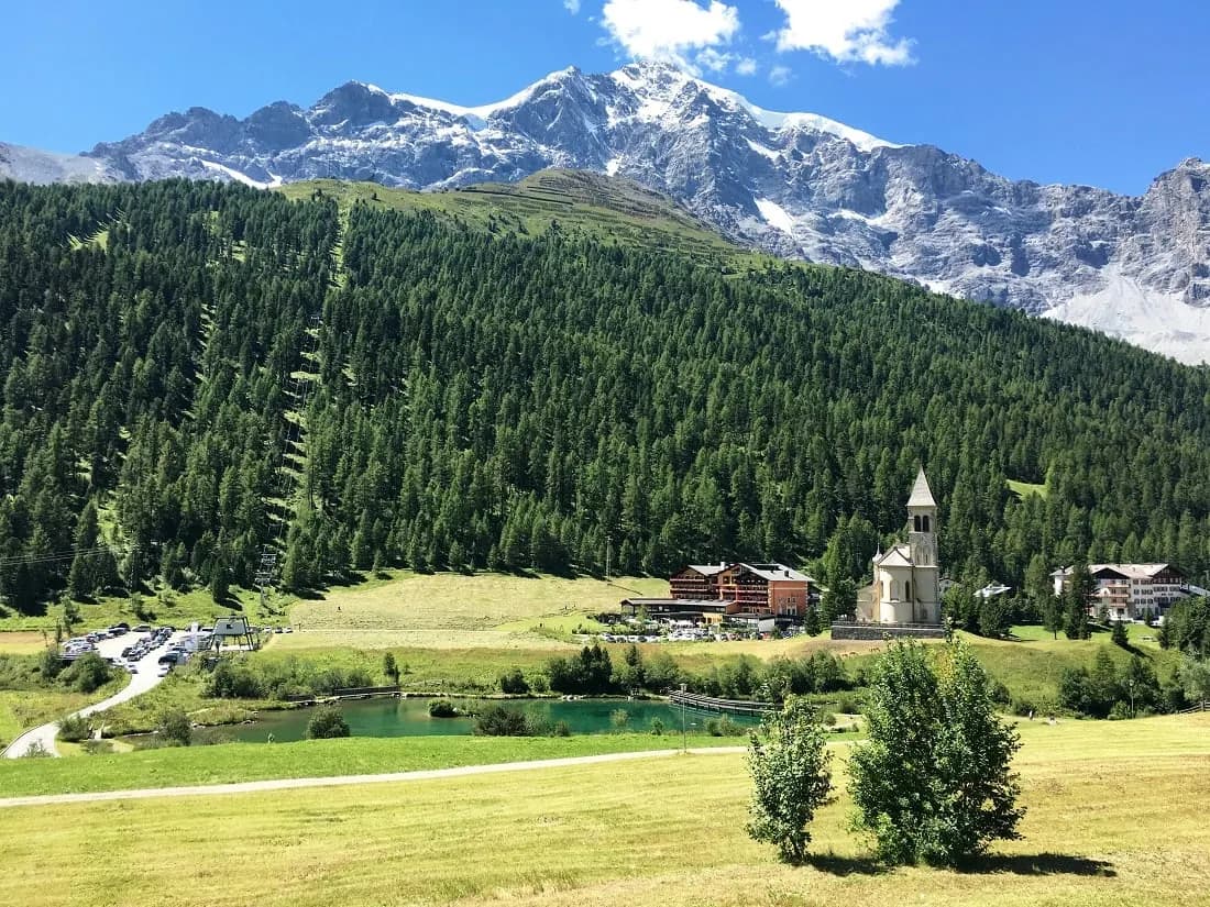 A small church and chalets in a green valley below forest and snowy peaks