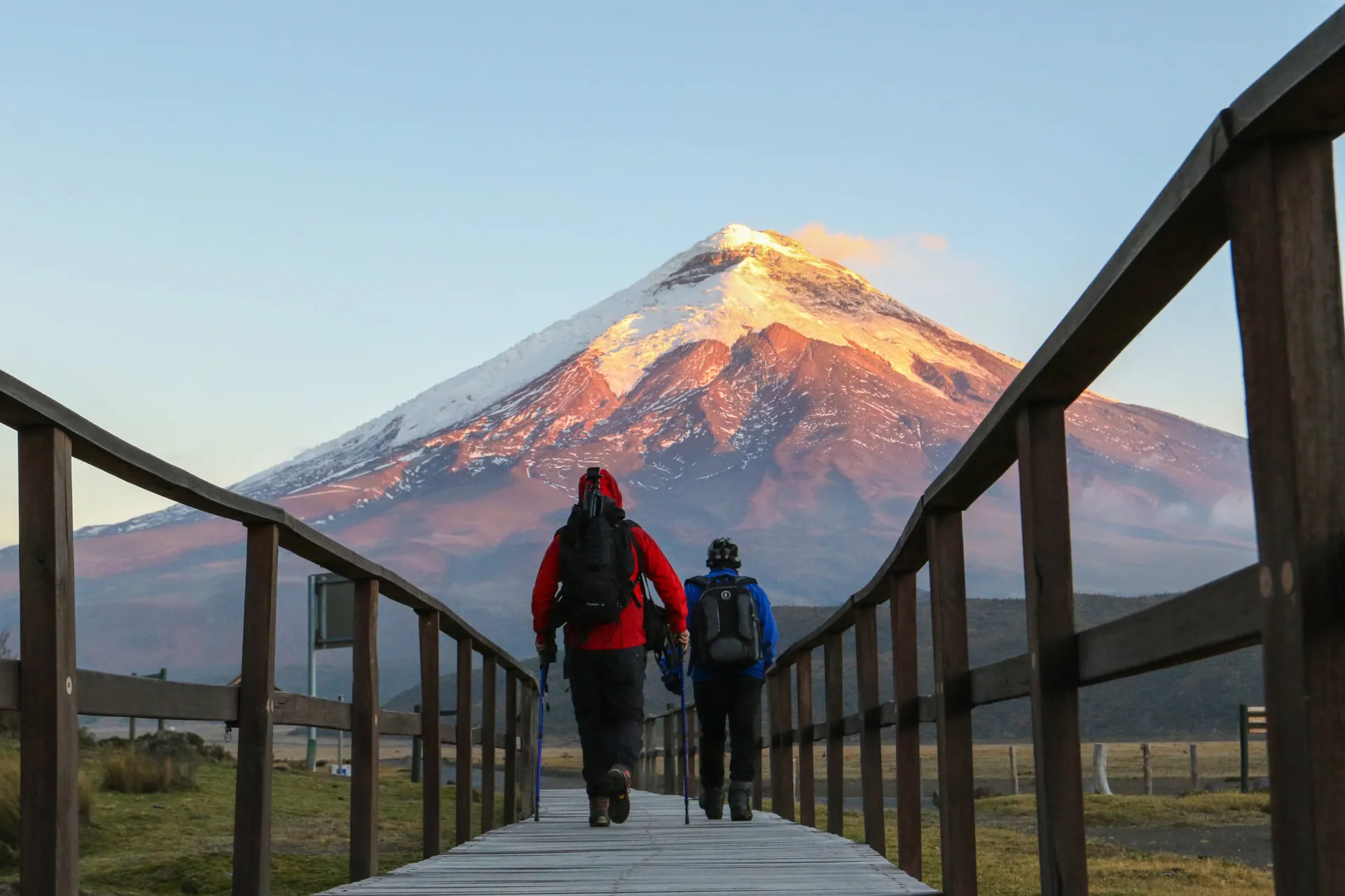 Two walkers on a boardwalk facing Cotopaxi glowing at dusk