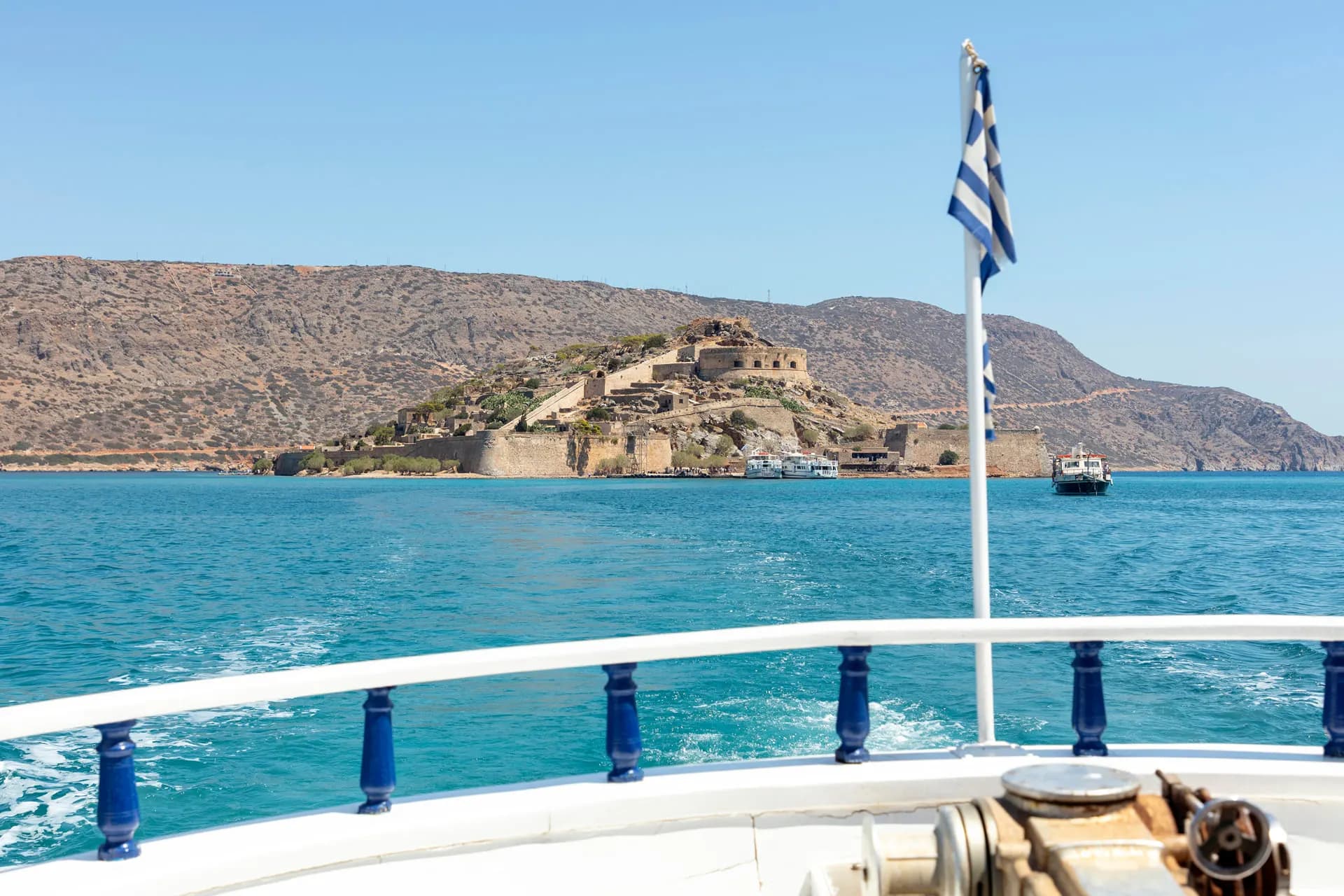 Spinalonga seen from the deck of a boat with a Greek flag