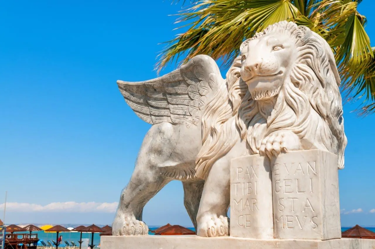 A white winged lion statue under a palm beside the beach