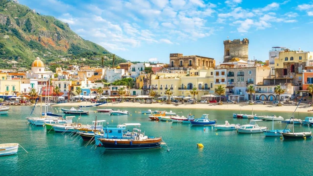 Fishing boats in a calm harbour below a town and a round stone tower on Ischia