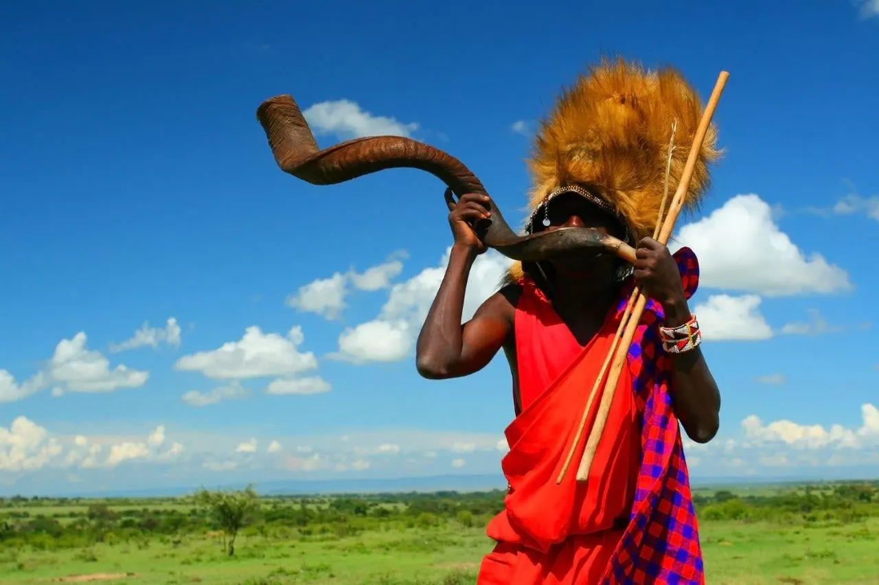 A Maasai man in a red shuka blowing a long horn under a blue sky