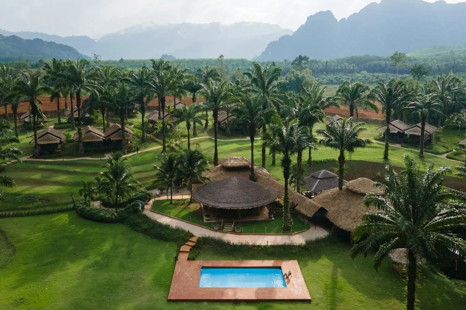 A camp with a small pool and thatched pavilion among palms, with limestone hills beyond