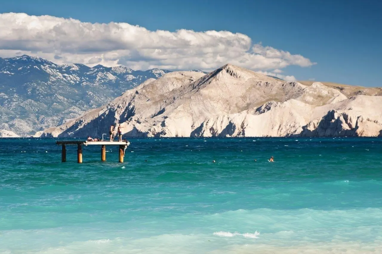 A pale beach and clear water below bare mountains on the island of Krk