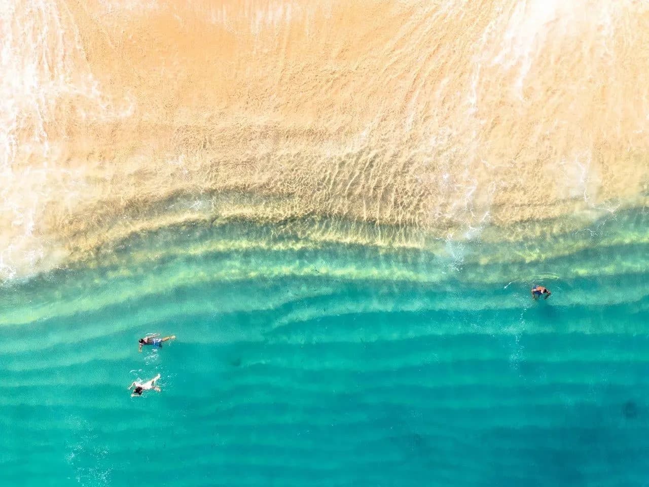 Swimmers in clear turquoise shallows off a sandy beach, seen from above