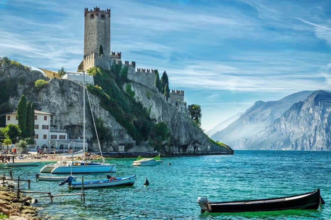 A castle tower on a rocky shore of Lake Garda, with boats in the clear water