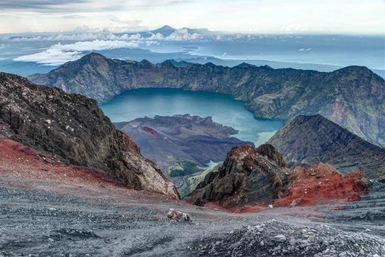 A blue crater lake inside a ring of volcanic peaks, seen from a high rocky rim