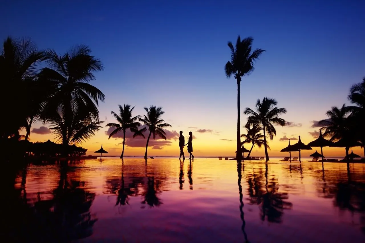 Palm trees and a couple in silhouette beside a still pool at sunset