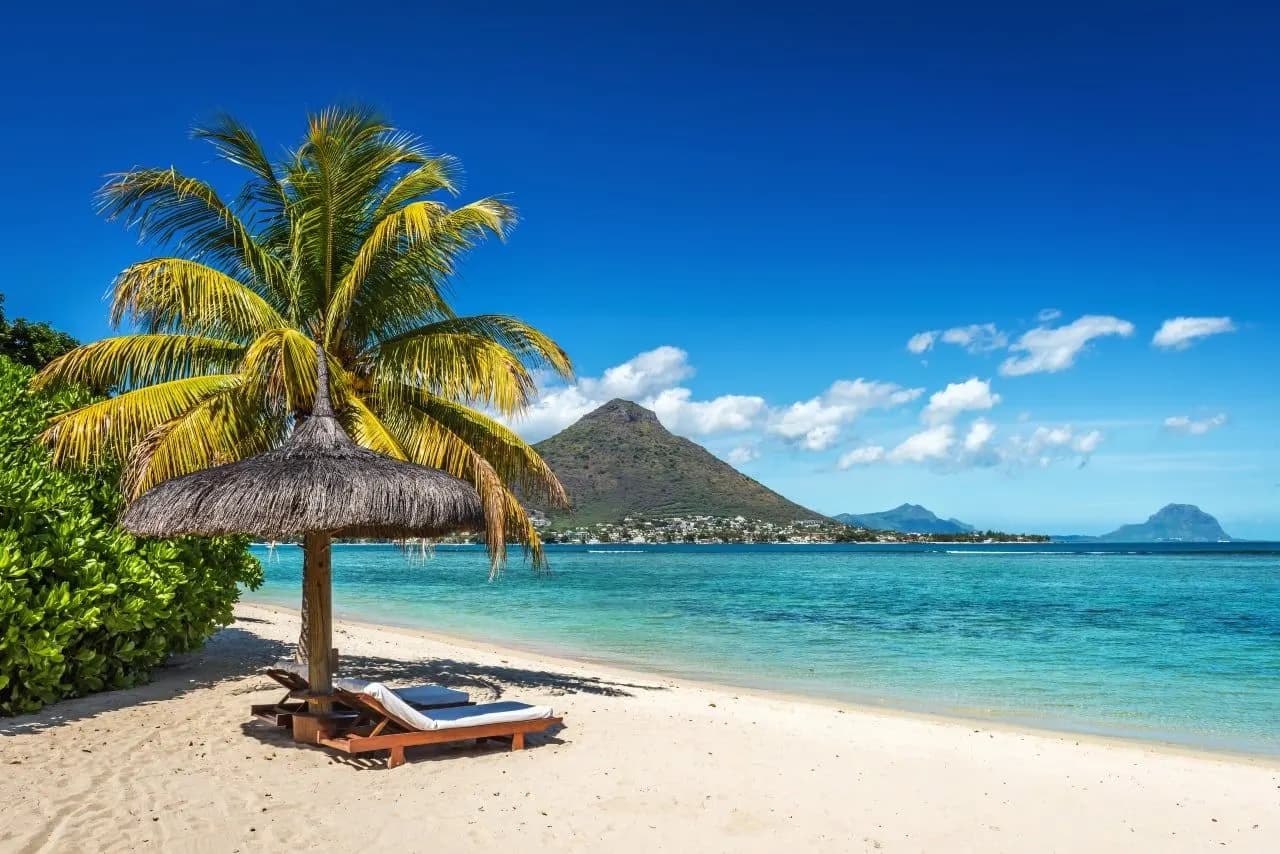 A thatched umbrella and sun lounger on a white beach, with a mountain across the bay