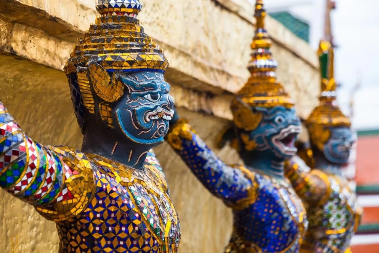 Mosaic guardian giants with blue faces supporting a golden chedi at the Grand Palace