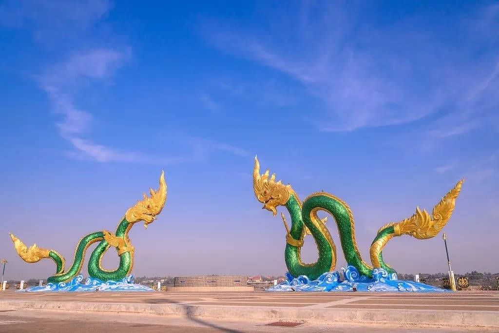 Green and gold naga sculptures rising from blue waves on a riverside square