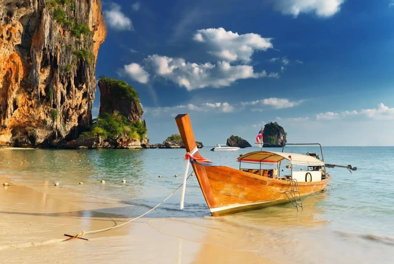 An orange long-tail boat pulled up on a beach beside towering limestone cliffs