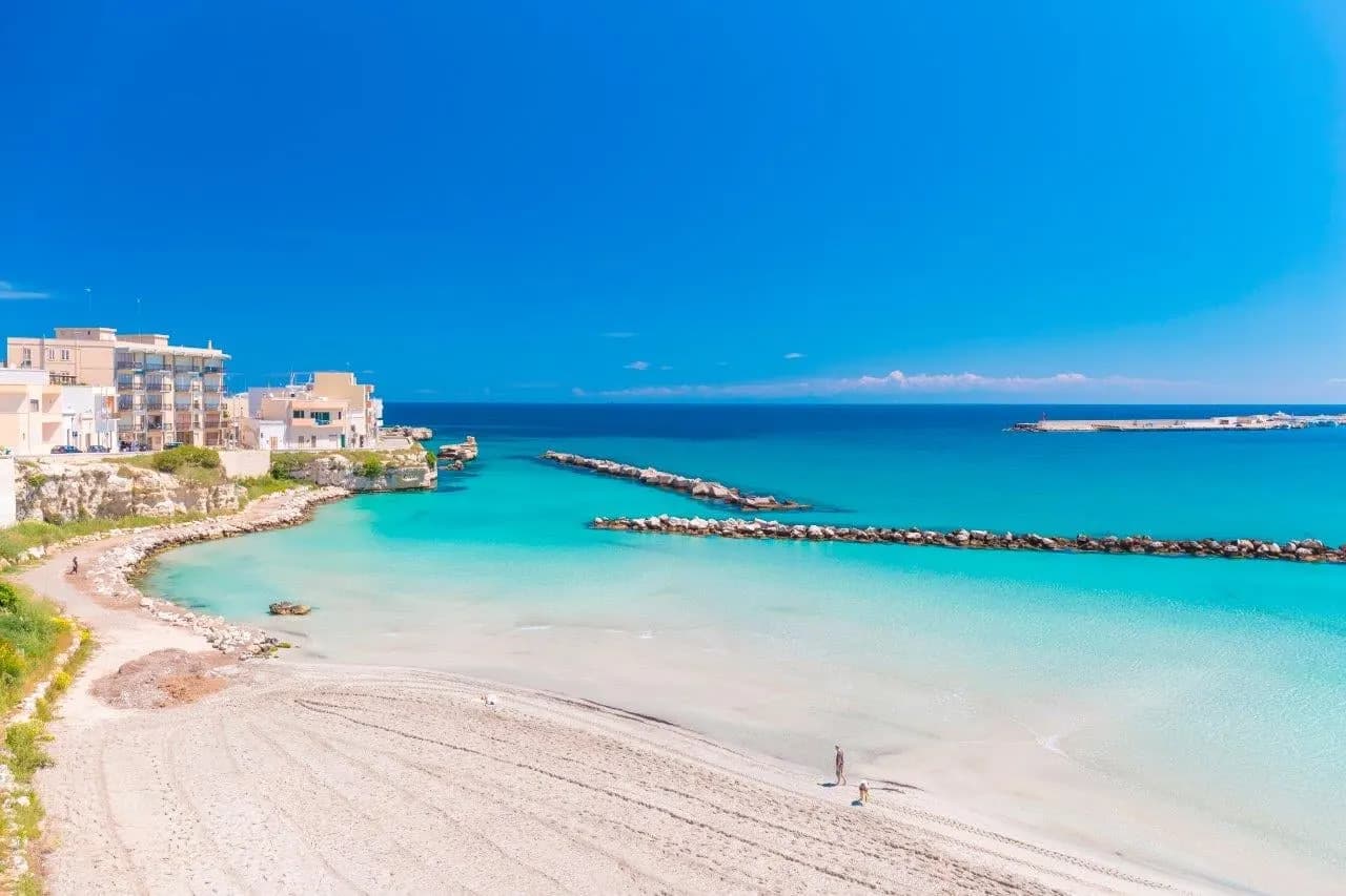 A pale sandy beach curving around turquoise water below a seaside town in Puglia