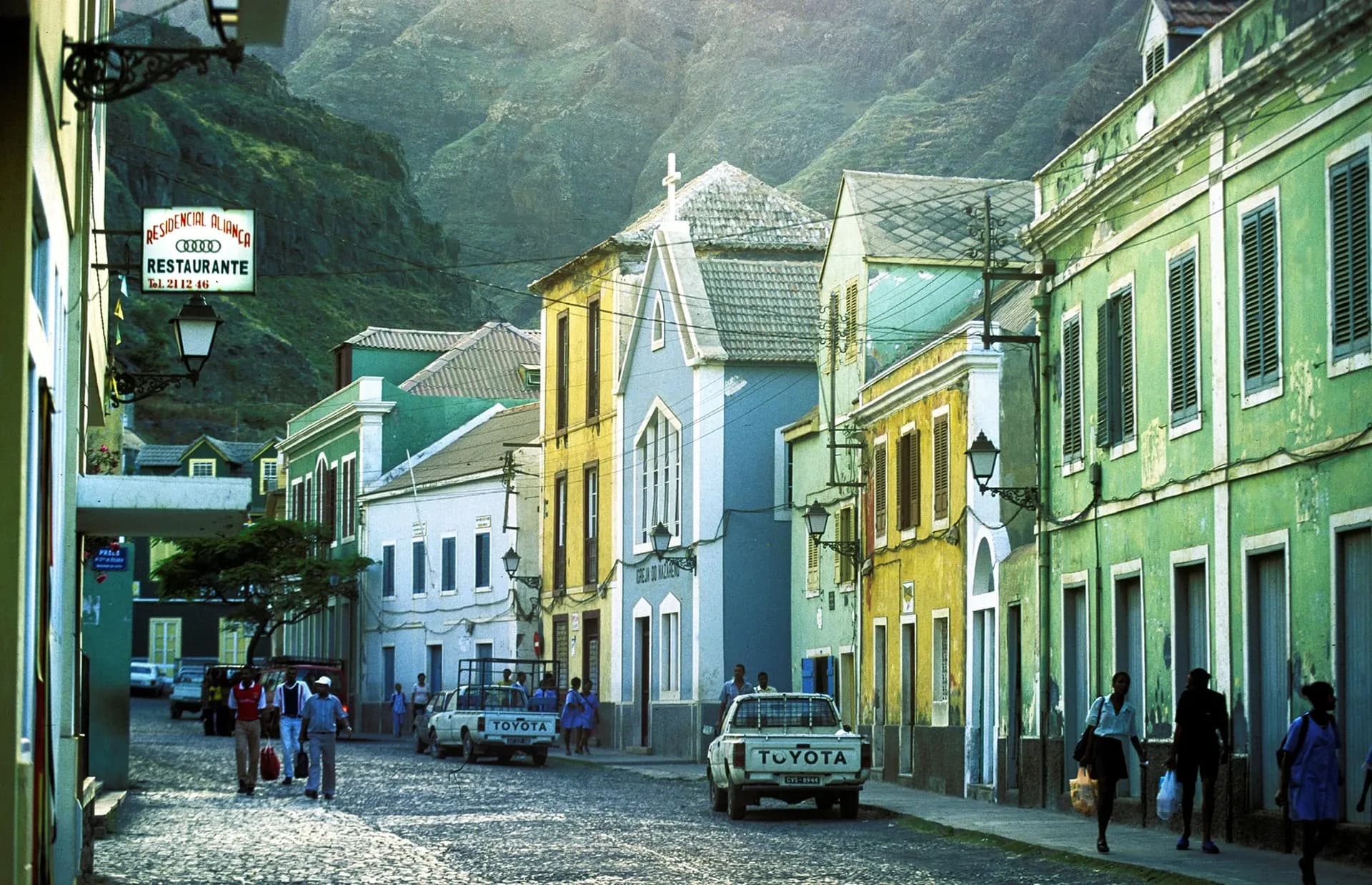 Pastel colonial buildings along a cobbled street below green mountains