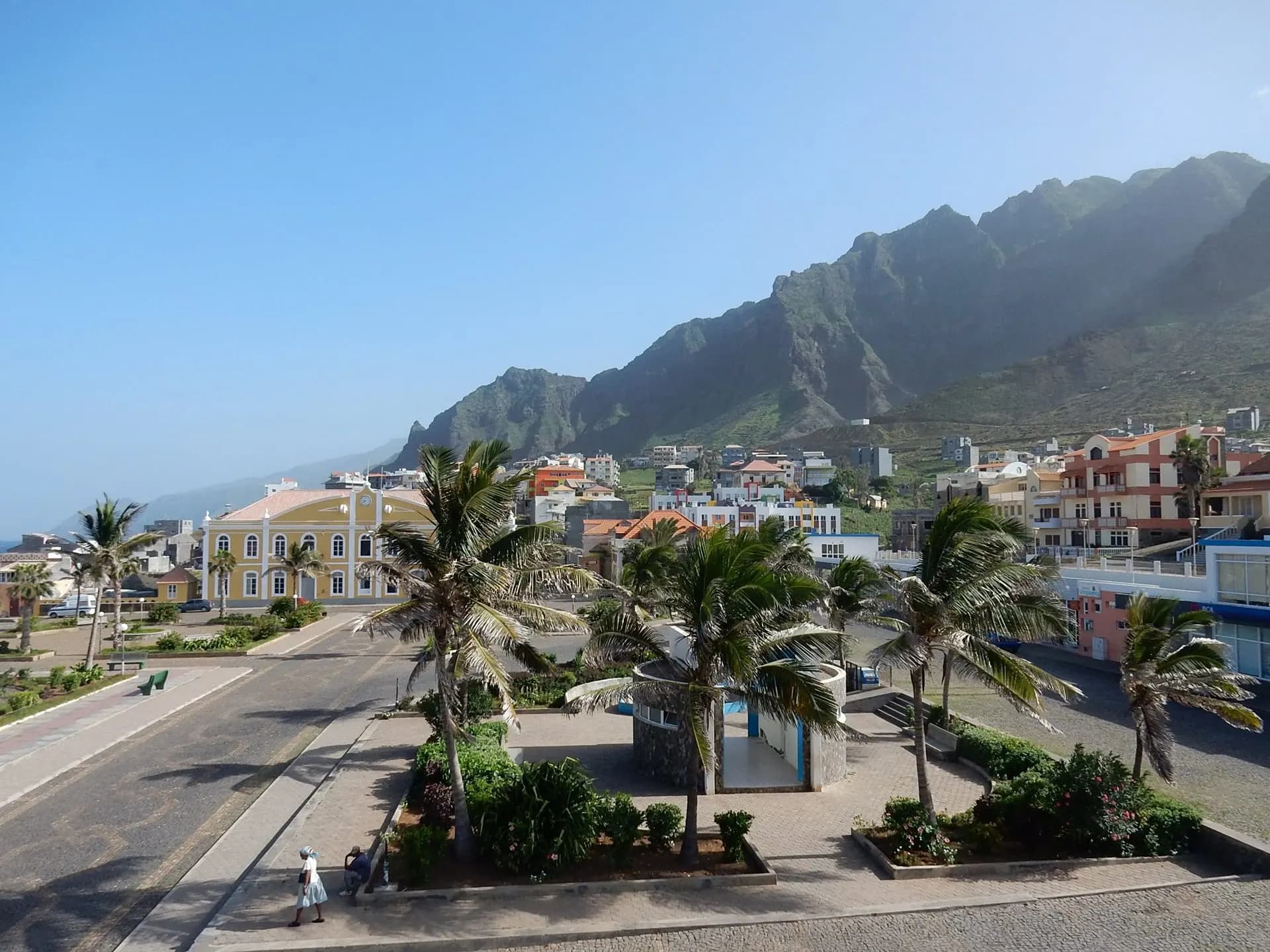 A palm-lined seafront road in a small town beneath mountains