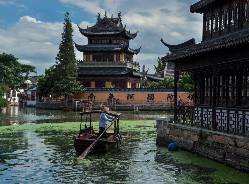 A boatman sculling a wooden boat along a canal past a pagoda in a water town