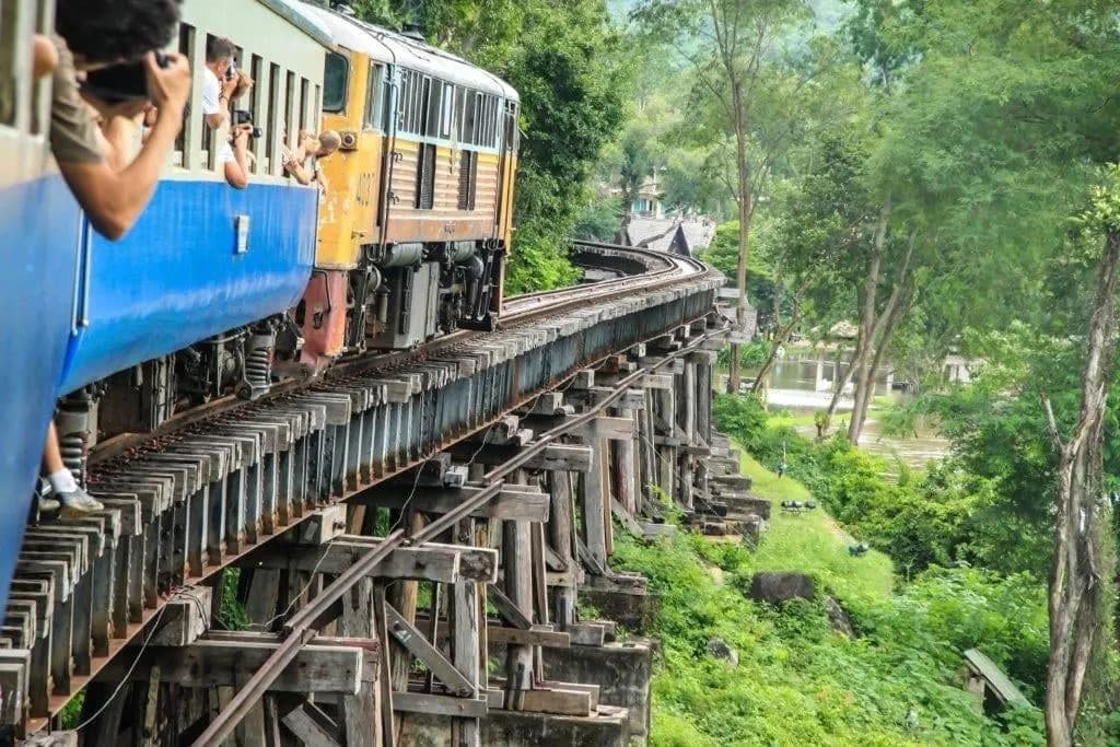 A blue train crossing a wooden railway viaduct beside the forest near Kanchanaburi