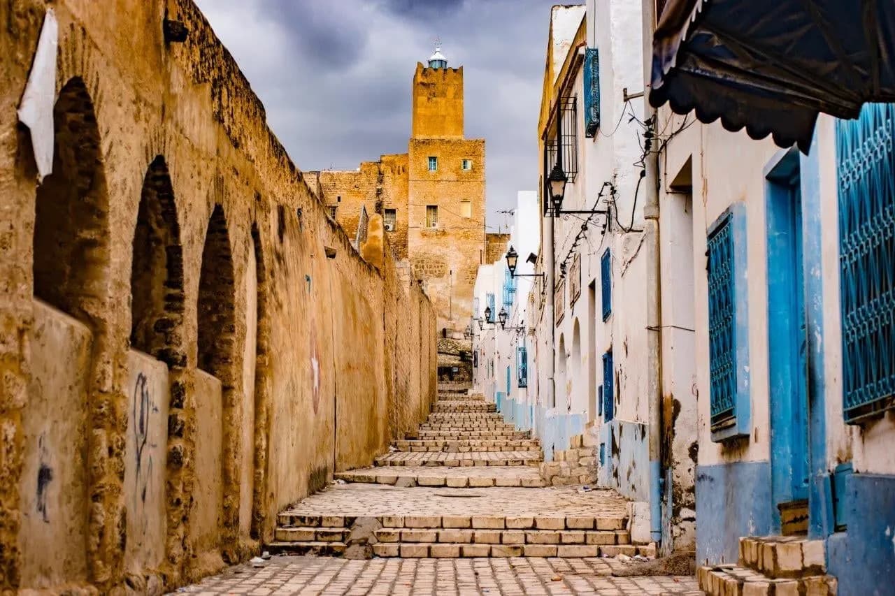 A stepped medina lane between old arched walls and blue doors, with a stone tower above
