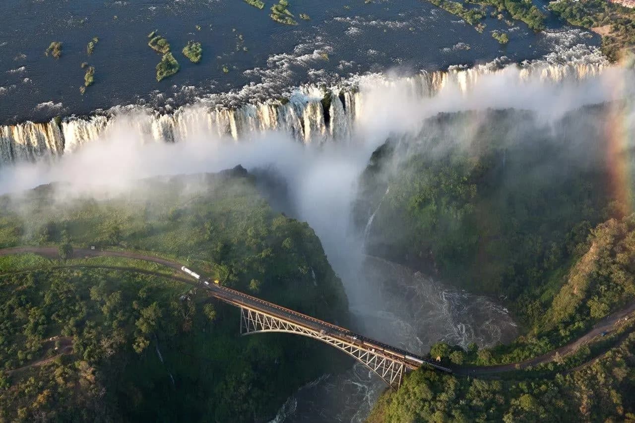 Spray rising from Victoria Falls above the gorge and the bridge, with a rainbow at its edge