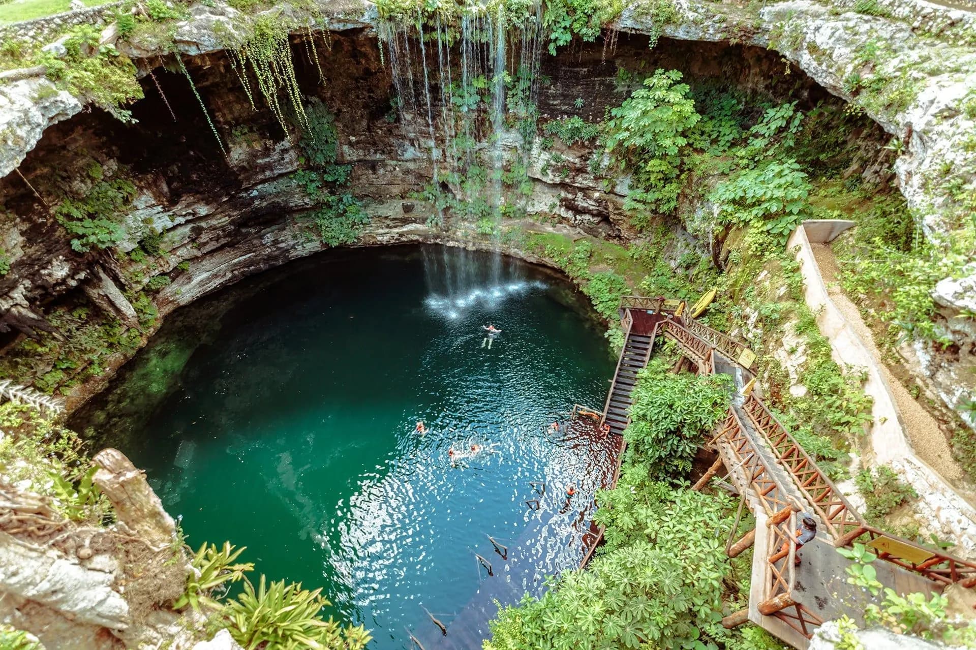 A cenote with clear green water at the bottom of a limestone sinkhole draped in vines