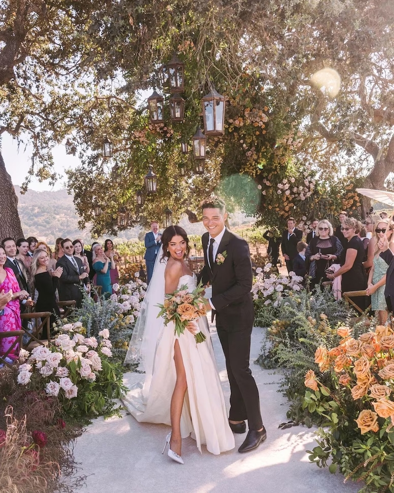 Happy bride and groom surrounded by vibrant wedding florals and guests