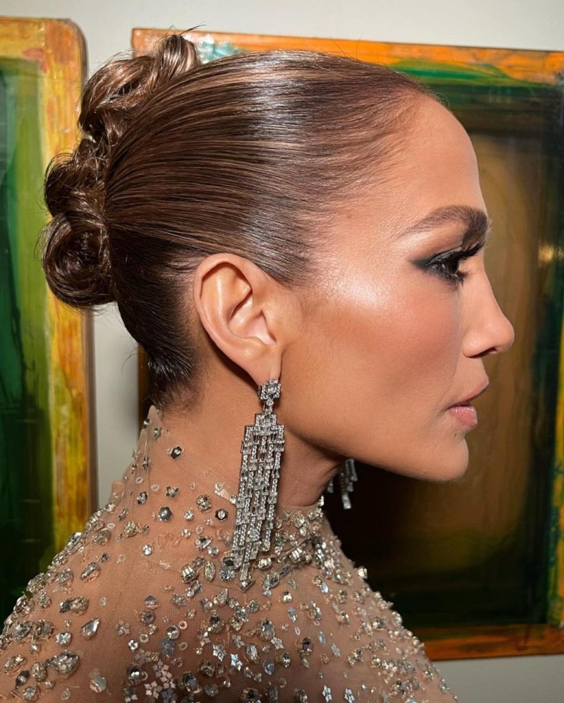 Jennifer Lopez on the red carpet at the Shotgun Wedding premiere, wearing silver earrings. Her hair styled in a sleek bun, with a radiant complexion and bold smoky eye makeup.