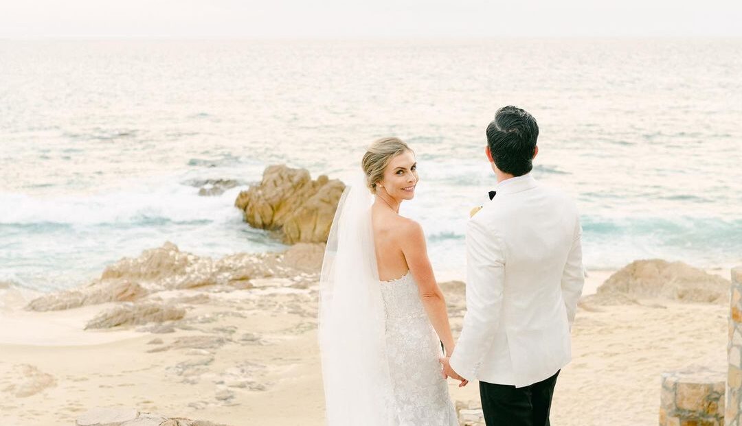 Bride and groom holding hands on a beach in Mexico, with ocean waves and rocky shoreline in the background, representing a romantic destination wedding.