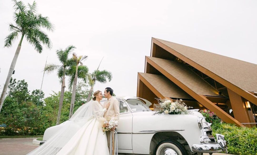 Bride and groom embracing in front of a classic white wedding car, surrounded by tropical palm trees and modern architecture, symbolizing a romantic destination wedding in Asia.