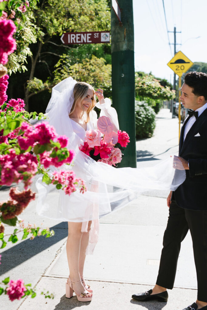 Bride in a white veil and mini dress holding vibrant pink bouquet, smiling at groom in tuxedo, surrounded by blooming flowers and street sign reading "IRENE ST," capturing the essence of Amy and Adam's intimate wedding celebration.