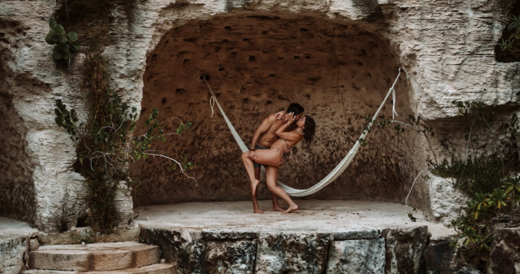 Couple embracing in a hammock within a natural stone alcove, surrounded by lush greenery, symbolizing romance and intimacy in a scenic Mexican wedding destination.