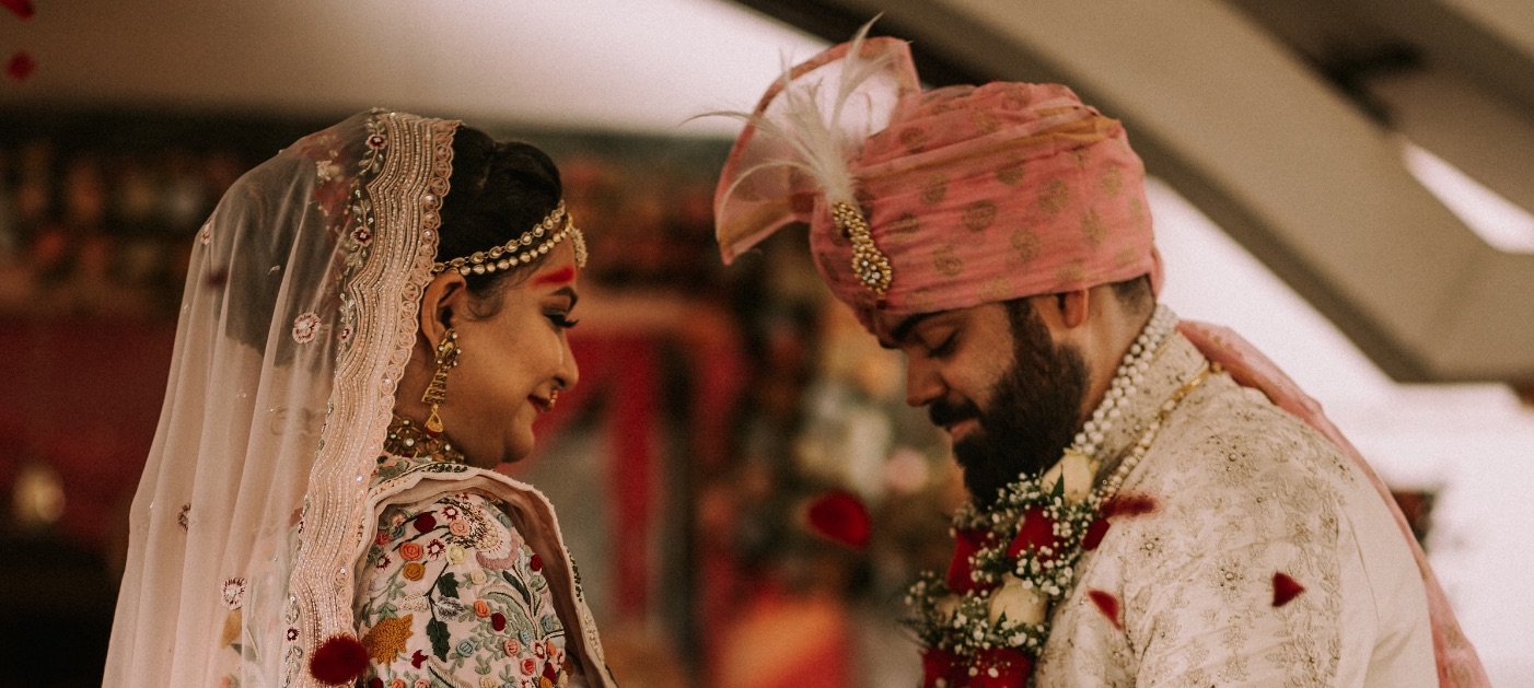 Bride and groom in traditional Asian wedding attire exchanging smiles, adorned with floral garlands, set against a richly decorated background, capturing the essence of cultural wedding celebrations.