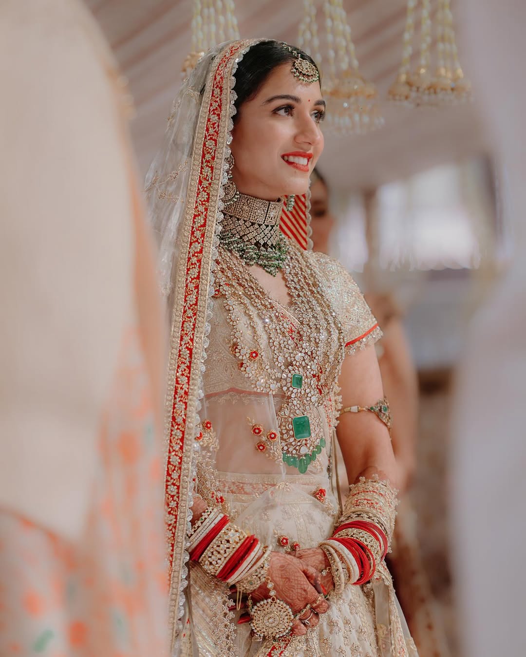 Bride in ornate traditional wedding attire, adorned with intricate jewelry and vibrant bangles, smiling gracefully during a cultural wedding ceremony.
