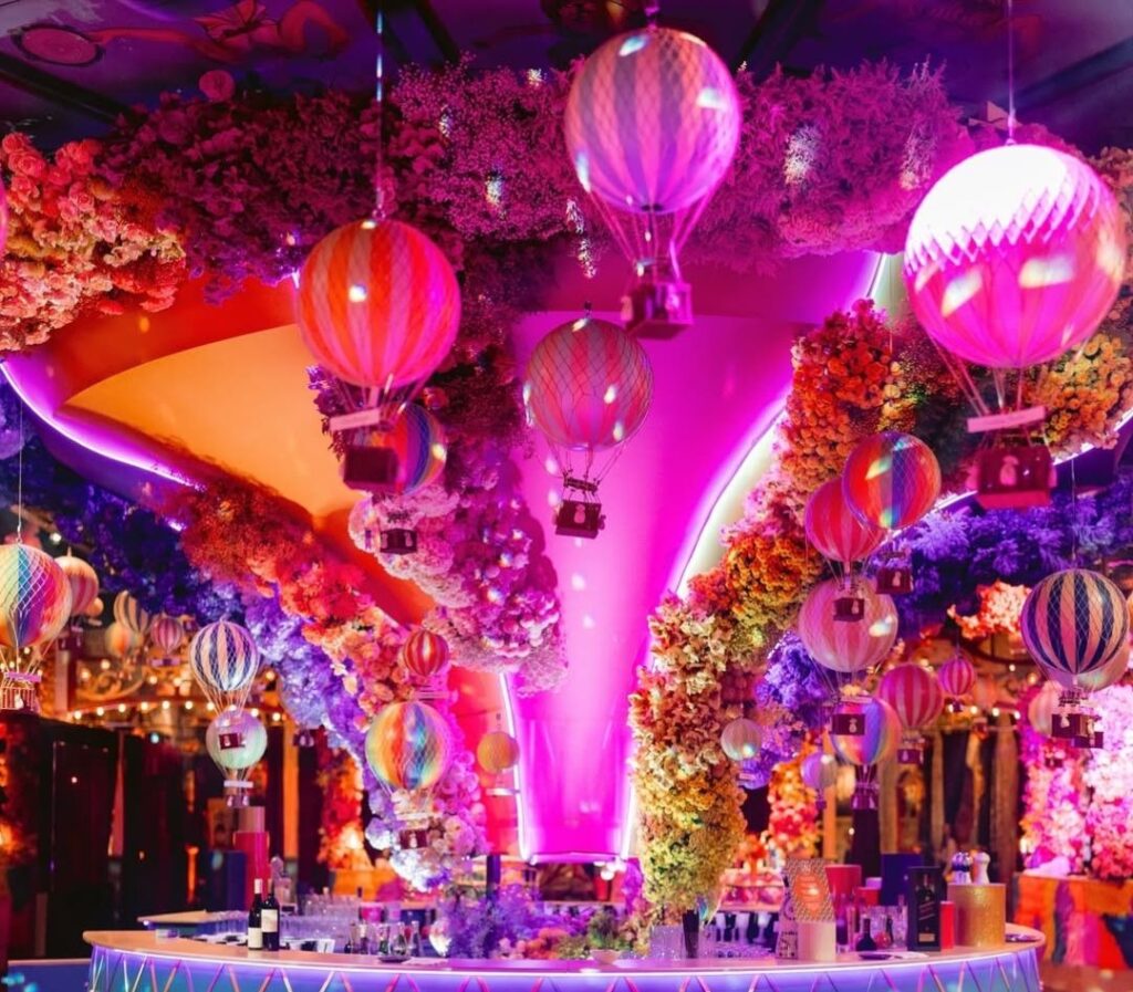 Colorful hot air balloons and vibrant floral decorations in a luxurious event space, showcasing extravagant themed wedding aesthetics by Alejandra Poupel.