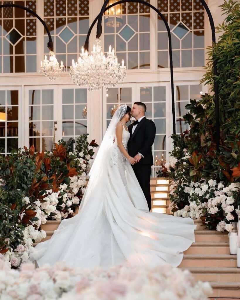 Where Sophistication Meets Storytelling: How Alysia Bridger brings love stories to life 7 Bride and groom embracing on elegant staircase adorned with floral arrangements, crystal chandelier above, capturing a moment of romance and luxury in wedding celebration.