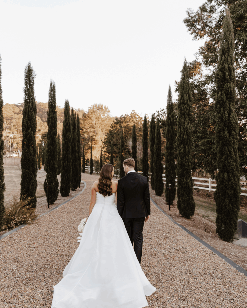 Where Sophistication Meets Storytelling: How Alysia Bridger brings love stories to life 1 Bride and groom walking hand in hand on a picturesque gravel path lined with tall cypress trees, embodying the elegance and sophistication of a luxury wedding planned by Alysia Bridger Events.