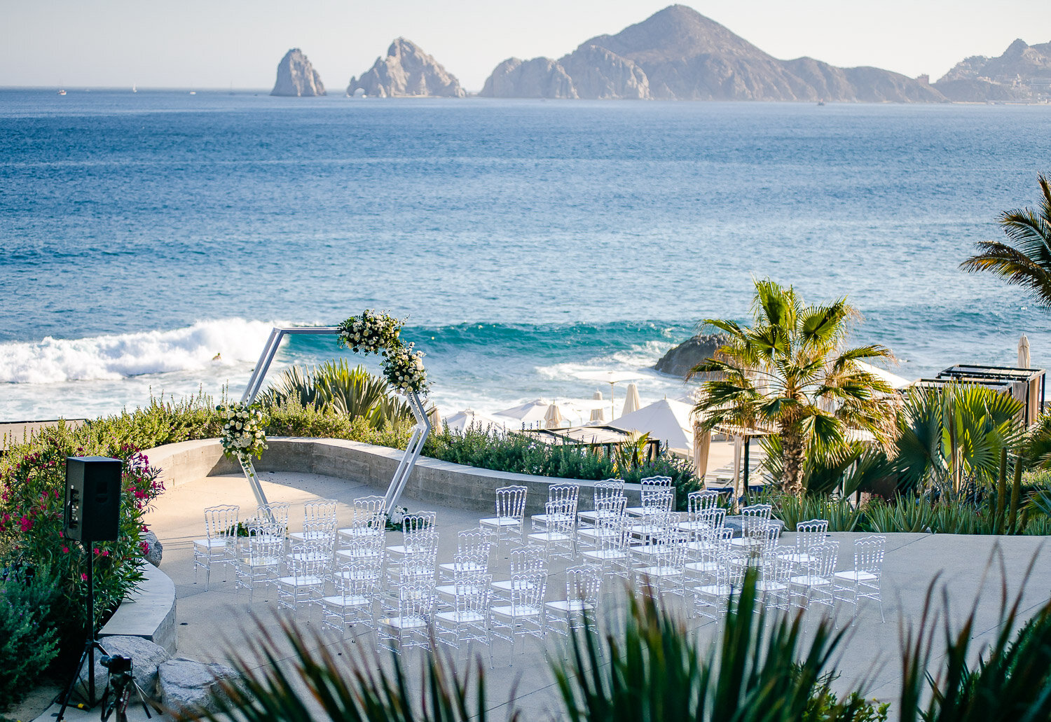 Beachside wedding ceremony setup in Los Cabos, featuring a floral arch, clear chairs, and ocean view with mountains in the background.
