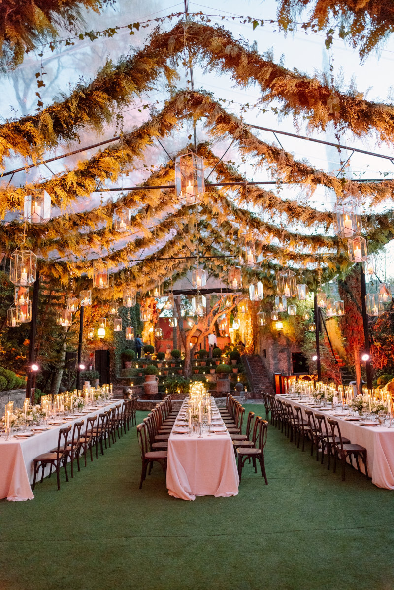 Elegant outdoor wedding reception setup in San Miguel de Allende, featuring long tables adorned with white linens, candles, and floral centerpieces, under a canopy of greenery and hanging lanterns.